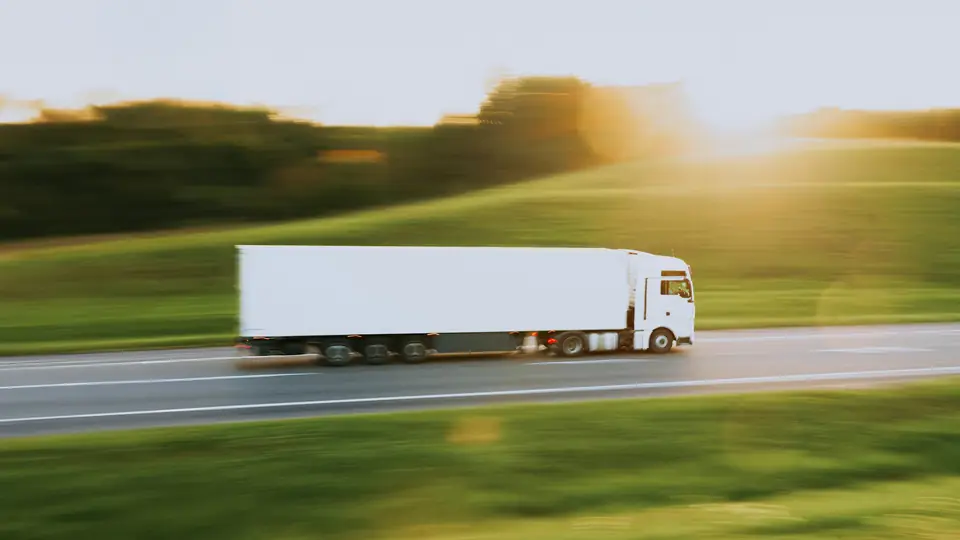 Vit lastbil med trailer på en motorväg genom gröna kullar i kvällssol